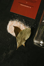 Load image into Gallery viewer, A close-up flat-lay of a pile of pink Himalayan salt crystals on a dark textured surface, with two dried bay leaves resting on top and part of a red branded card visible at the top edge.

