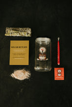 Load image into Gallery viewer, A flat-lay of a Meraki Solar Return Spell Kit on a dark textured background, including a sage and sweetgrass bundle, a gold instruction card, a clear glass jar with an illustration, a red soy candle, salt and crystal, and a small red matchbook.
