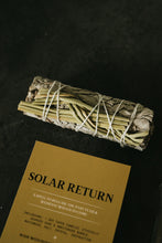 Load image into Gallery viewer, A sage and sweetgrass bundle on a flat black surface above a gold instruction card titled "Solar Return."
