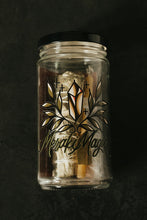 Load image into Gallery viewer, A clear glass jar with black lid against a black textured background. The jar has a black and gold toned graphic of a crystal and the words "Meraki Magic." In the jar is one soy taper candle, yerba santa and sage bundle, bay leaf, salt, lavender, and instruction card.
