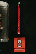 Load image into Gallery viewer, Flat-lay product shot of a red soy taper candle, above a red matchbook, and a partial view of a glass jar with a graphic on it, all on a black background.
