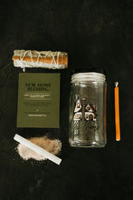 Load image into Gallery viewer, A New Home Blessing Spell Kit arranged on a dark textured background, including a sage bundle with cinnamon stick, a green instruction card with 'New Home Blessing' text, a clear glass jar with a house illustration and 'Blessing For Your New Home' text, a selenite wand on a pile of salt, and a soy taper candle.
