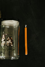 Load image into Gallery viewer, A clear glass jar candle with a black lid and printed house graphic, shown next to a soy taper candle on a dark textured background.

