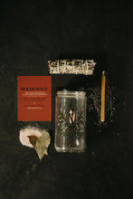 Load image into Gallery viewer, A flat-lay product presentation of the Meraki Magic Spell Kit on a dark textured background, featuring a clear glass jar with a black lid and graphic art, a sage bundle, a soy candle, bay leaves and salt, with instruction card.
