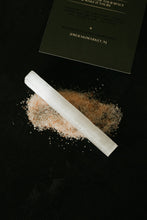 Load image into Gallery viewer, A selenite crystal wand resting on a small pile of salt crystals, with a dark green branded card partially visible in the background on a black surface.
