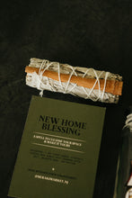 Load image into Gallery viewer, A sage bundle and cinnamon stick wrapped in white string rests on top of an olive green instruction card titled 'NEW HOME BLESSING' against a dark textured background.
