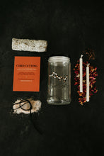 Load image into Gallery viewer, A cord cutting kit arranged on a dark textured background, including a sage bundle, an orange instruction card labeled CORD CUTTING, a clear glass jar with a black lid, two white taper candles, rose petals, clove, a cord on top of a small pile of salt, and sage bundle.

