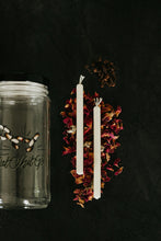 Load image into Gallery viewer, Two white taper candles arranged vertically on a bed of rose petals, some clove above them, and a clear glass jar partially visible on the left, all set against a dark black background.
