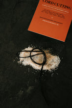 Load image into Gallery viewer, An orange instruction card titled 'Cord Cutting' lies on a dark textured surface next to a small pile of white salt and a black cord tied in a loop.

