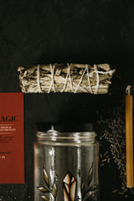 Load image into Gallery viewer, A sage smudge bundle tied with white string, displayed on a dark surface alongside a red card, a clear glass jar with a black lid and crystal illustration, and a yellow candle.
