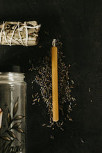 Load image into Gallery viewer, A single yellow taper candle lies on a dark surface, surrounded by scattered dried lavender buds. A white sage bundle tied with string is partly visible at the top left, and the edge of a glass jar with botanical artwork is visible at the bottom left.
